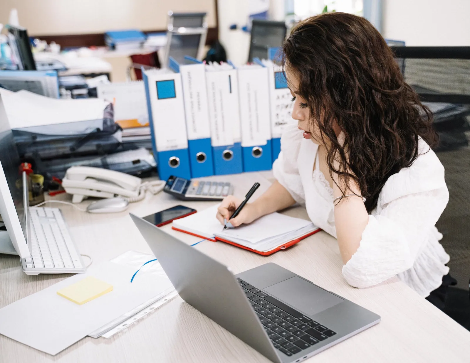 An office worker writes notes by hand beside her laptop and a row of blue ring binders