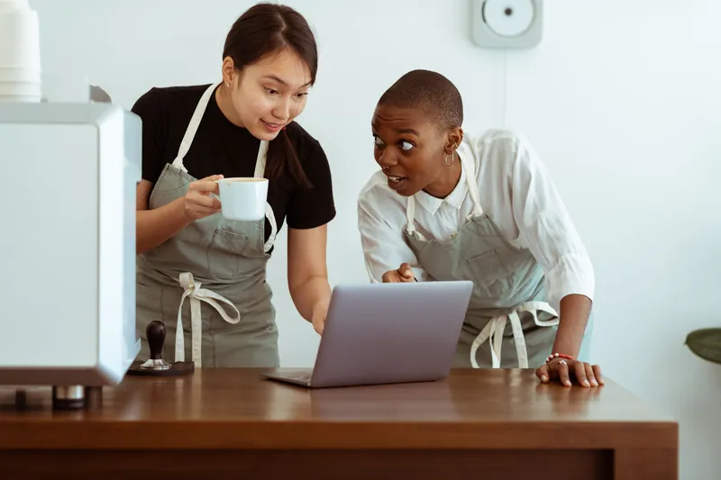 Two café staff in aprons look at a laptop together on the counter during a quiet moment