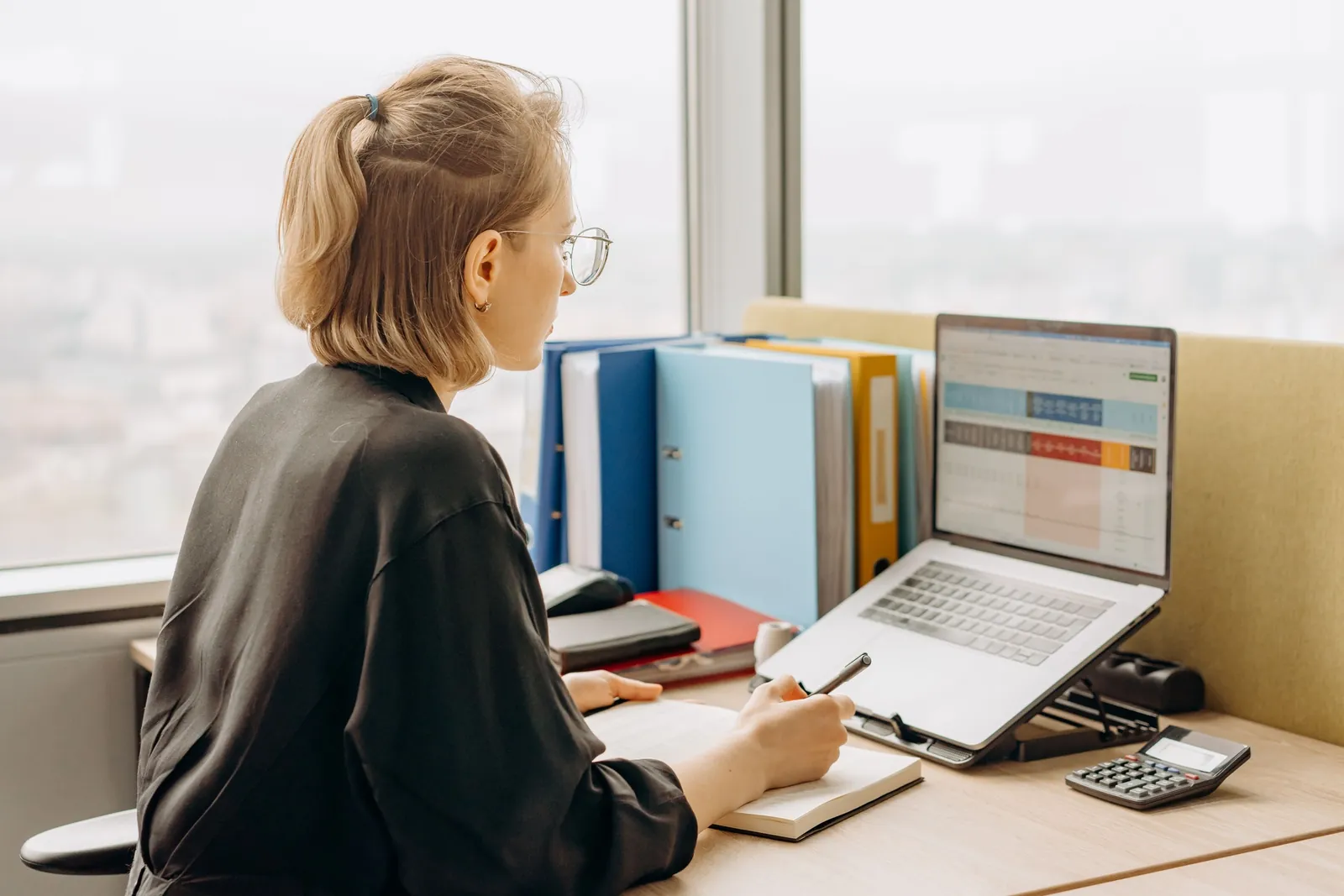 A woman works through a spreadsheet on a laptop at a desk by a window, with ring binders behind her