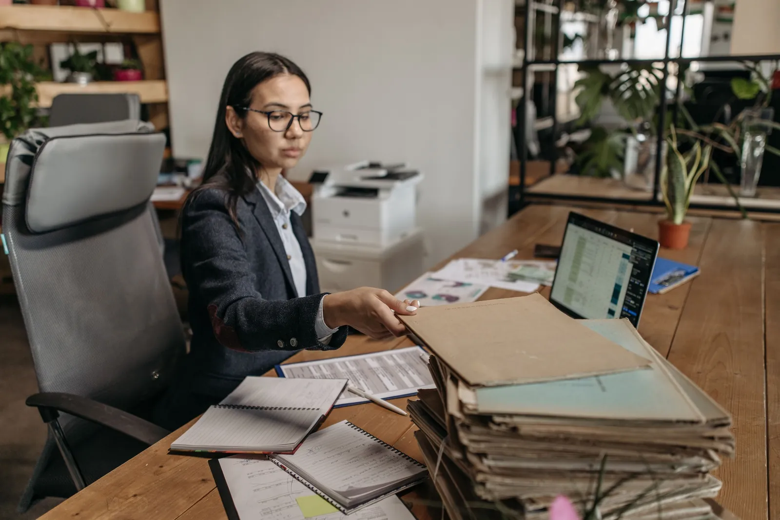 A woman at a wooden desk works through a pile of paper files beside her laptop