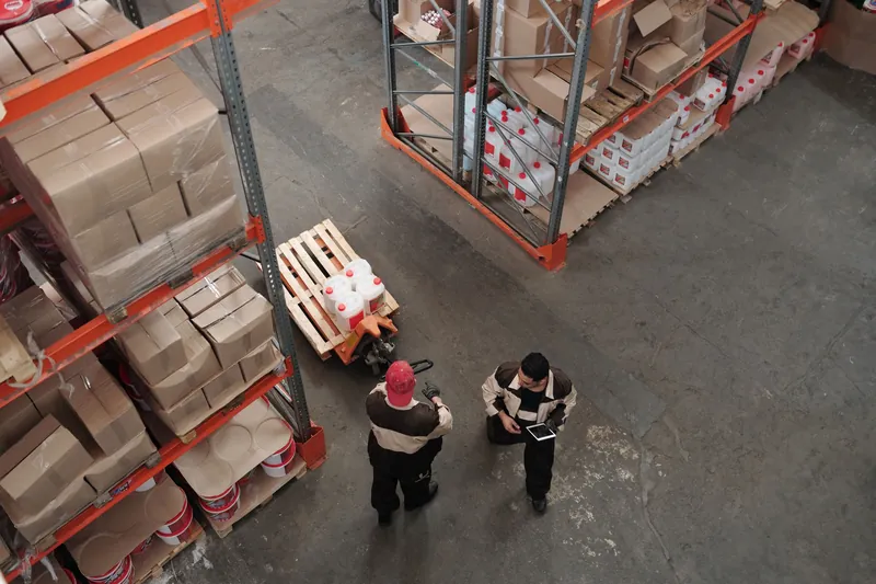 Seen from above, two warehouse workers check a tablet beside a pallet truck between tall shelves of boxes